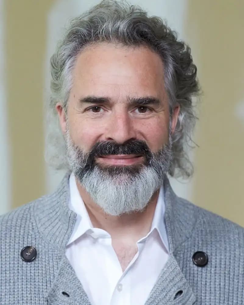 Portrait of a man with gray hair and beard wearing a gray sweater and white shirt, smiling at the camera